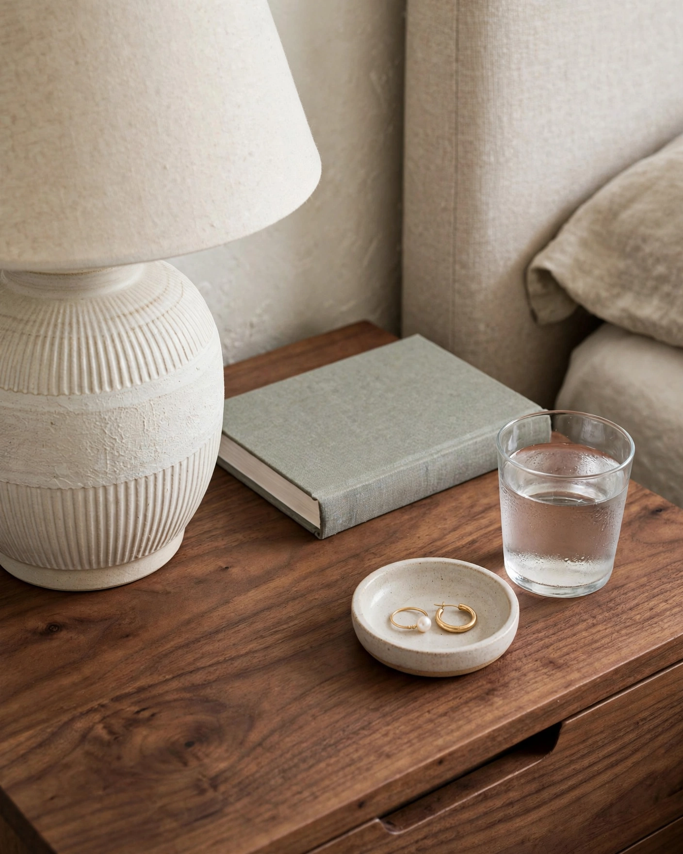 A tidy walnut nightstand holds a ceramic lamp, book, jewelry dish, and drinking glass.